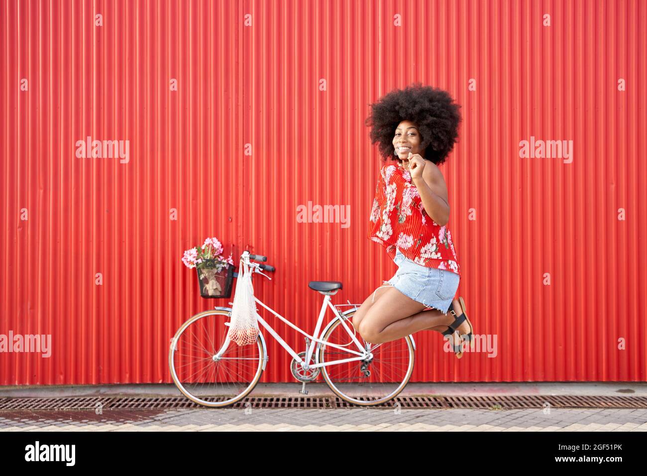 Glückliche Afro-Frau, die mit dem Fahrrad vor der roten Wand springt Stockfoto