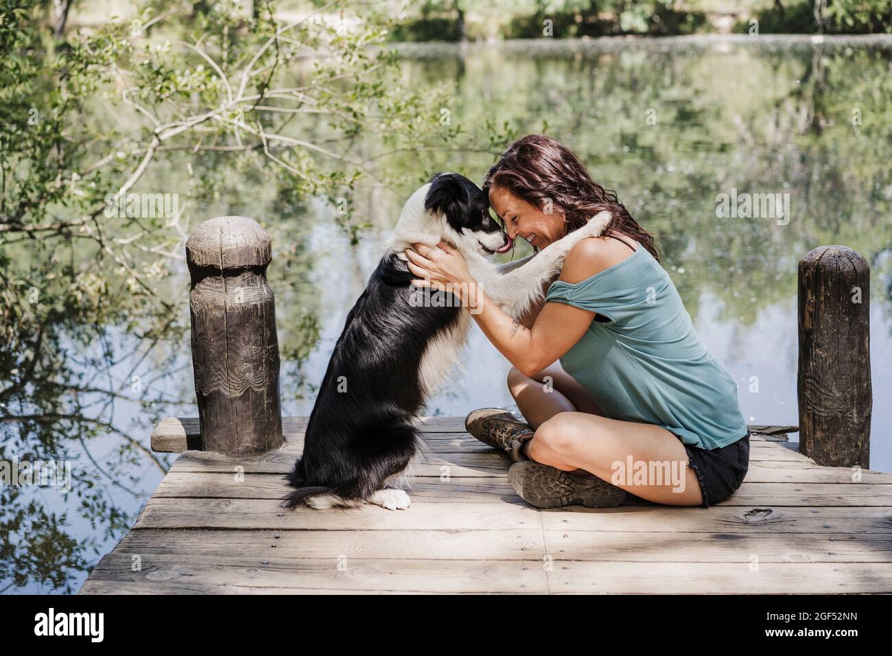 Lächelnde Frau, die Hund umarmt, während sie am Pier am See im Wald sitzt Stockfoto