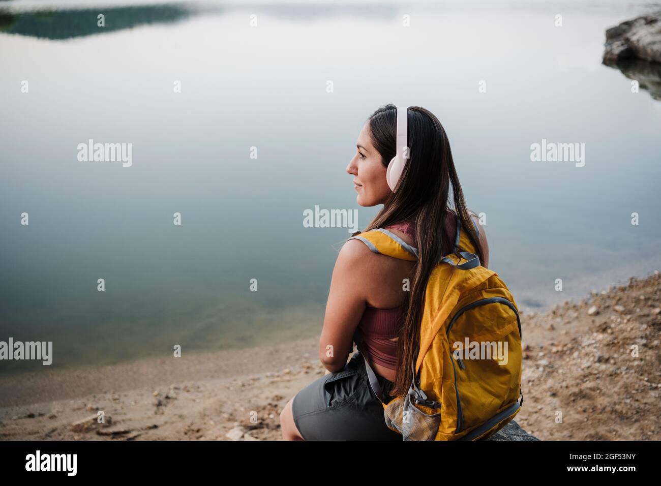 Junge Backpacker-Frau schaut weg, während sie auf Felsen am Seeufer sitzt Stockfoto