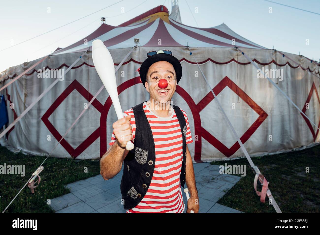 Lächelnder männlicher Clown, der die Jongliernadel hält, während er vor dem Zirkuszelt steht Stockfoto