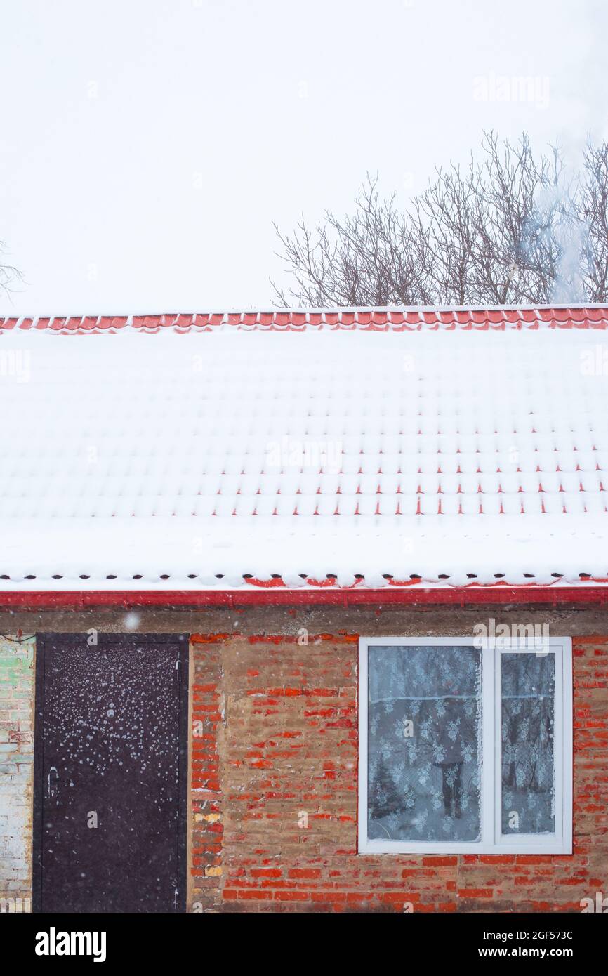 Ein Haus mit einem Metalldach, das mit fallendem Schnee bedeckt ist. Wintersaison im Dorf. Stockfoto