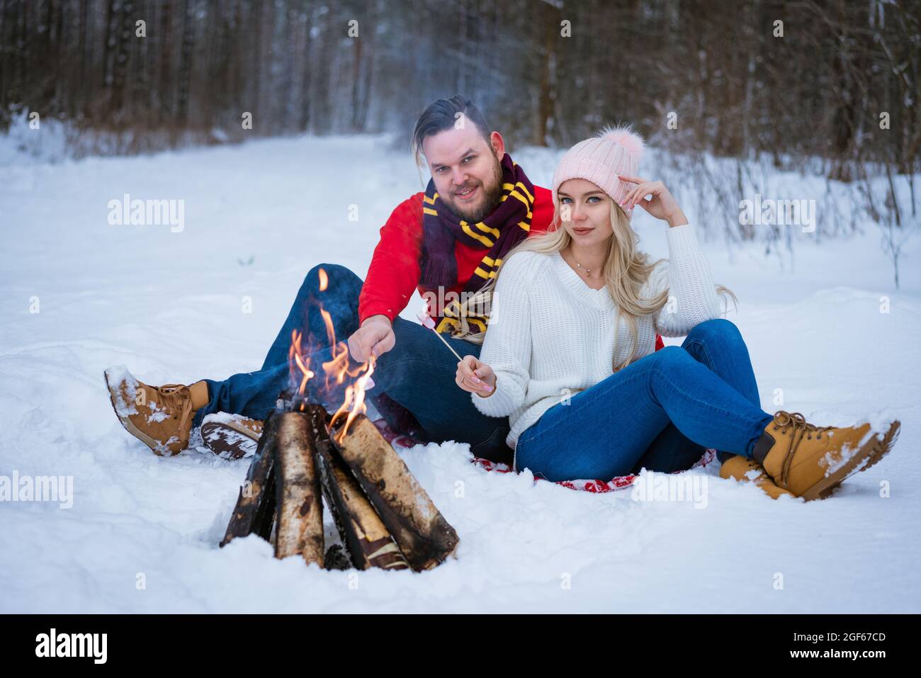 Im Winter sitzen ein junges Paar, ein erwachsener Mann und eine Frau im Schnee in der Nähe eines Lagerfeuers. Junge Menschen tragen Jeans, Pullover und Winterschuhe. Winter bewölkt Tag, Bäume im Hintergrund. Stockfoto