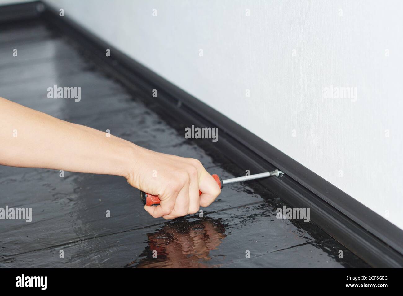 Ein Handwerker installiert eine schwarze PVC-Sockelleiste aus Kunststoff, um Kabel mit einem Schraubendreher an einer weißen Wand zu verbergen, während die Wohnung renoviert wird Stockfoto