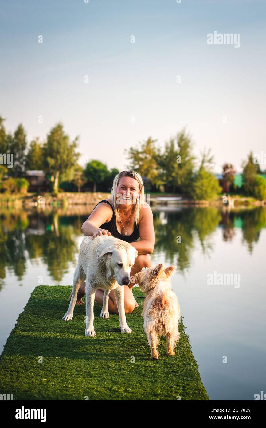 Fröhliche lachende Frau hat Spaß mit ihren Hunden am Pier am See an sonnigen Tagen im Sommer. Labrador Retriever und Cairn Terrier mit Haustierbesitzer im Freien Stockfoto