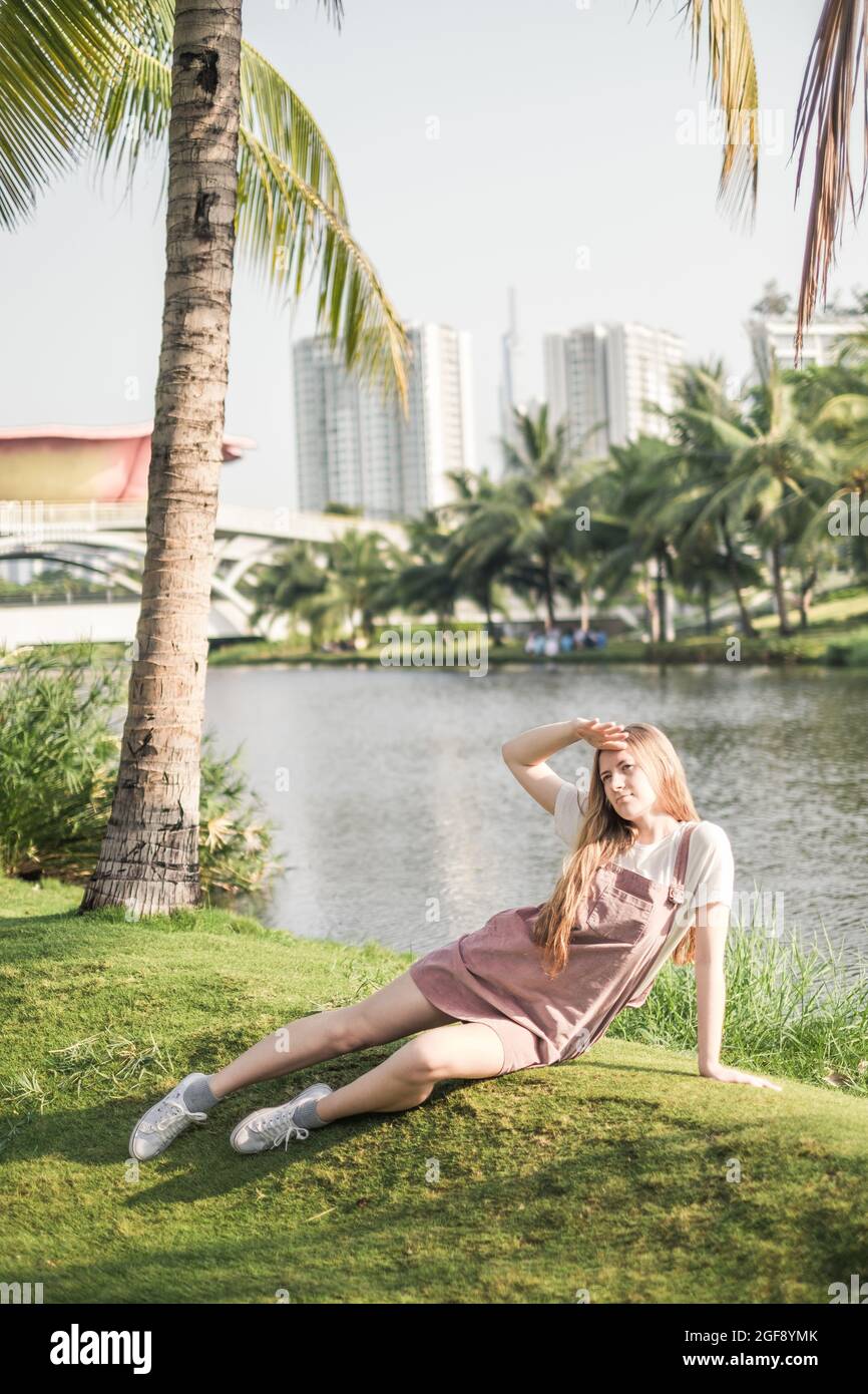 Hübsches Mädchen in legerer Kleidung, das im Park auf dem Rasen sitzt. Lange blonde Haare. Modisches, lässiges Sommer- oder Frühlingsoutfit Stockfoto