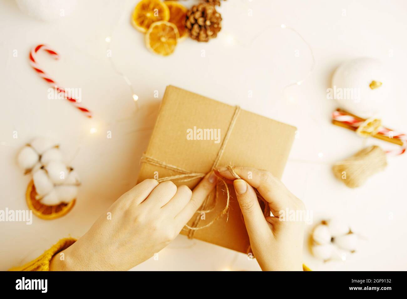 Verpackung von Weihnachten Geschenkbox. Tannenzapfen, trockene Orangenscheiben und andere Dekorationen auf dem Tisch. Mädchen im Pullover bereitet Überraschung für das neue Jahr. Festliche Atmosphäre. Damenhänden binden Band im Knoten. Stockfoto