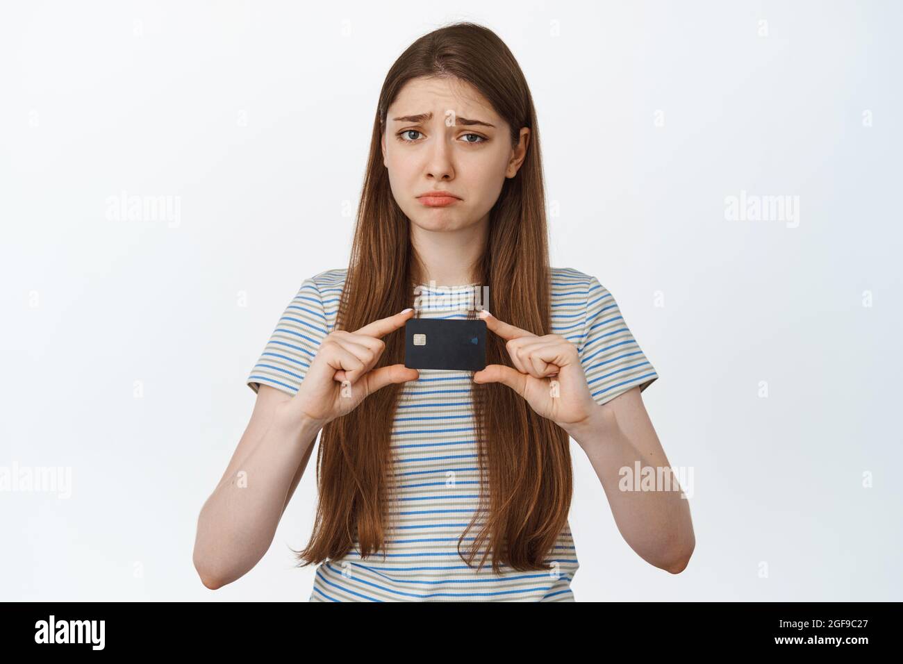 Traurige Frau zeigt Kreditkarte, Pouts und Stirnrunzeln enttäuscht, steht vor weißem Hintergrund Stockfoto