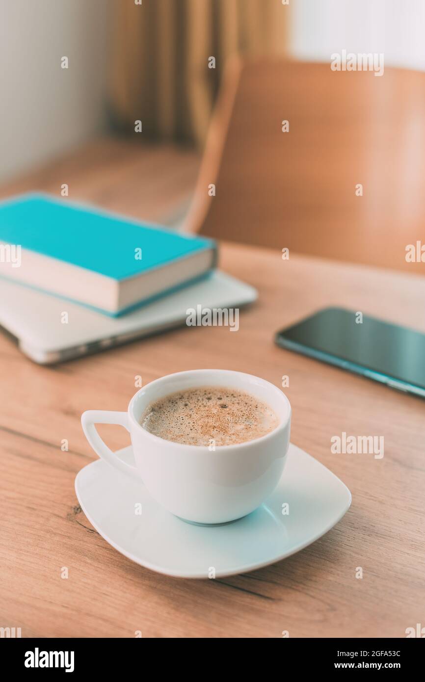 Bedeutung von Koffein und Kaffee für die freiberufliche Arbeit. Kaffeetasse, mobiles Smartphone, Laptop-Computer und ein Buch auf dem heimischen Bürotisch. Stockfoto