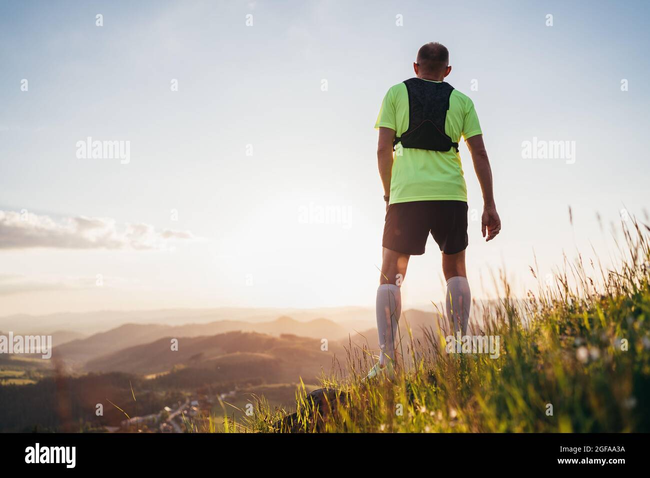 Rückansicht des Active Mountain Trail Läufers gekleidet helles T-Shirt mit Rucksack genießen den Sonnenuntergang, während er Himmel läuft durch malerische Hügel bei Sonnenuntergang Stockfoto