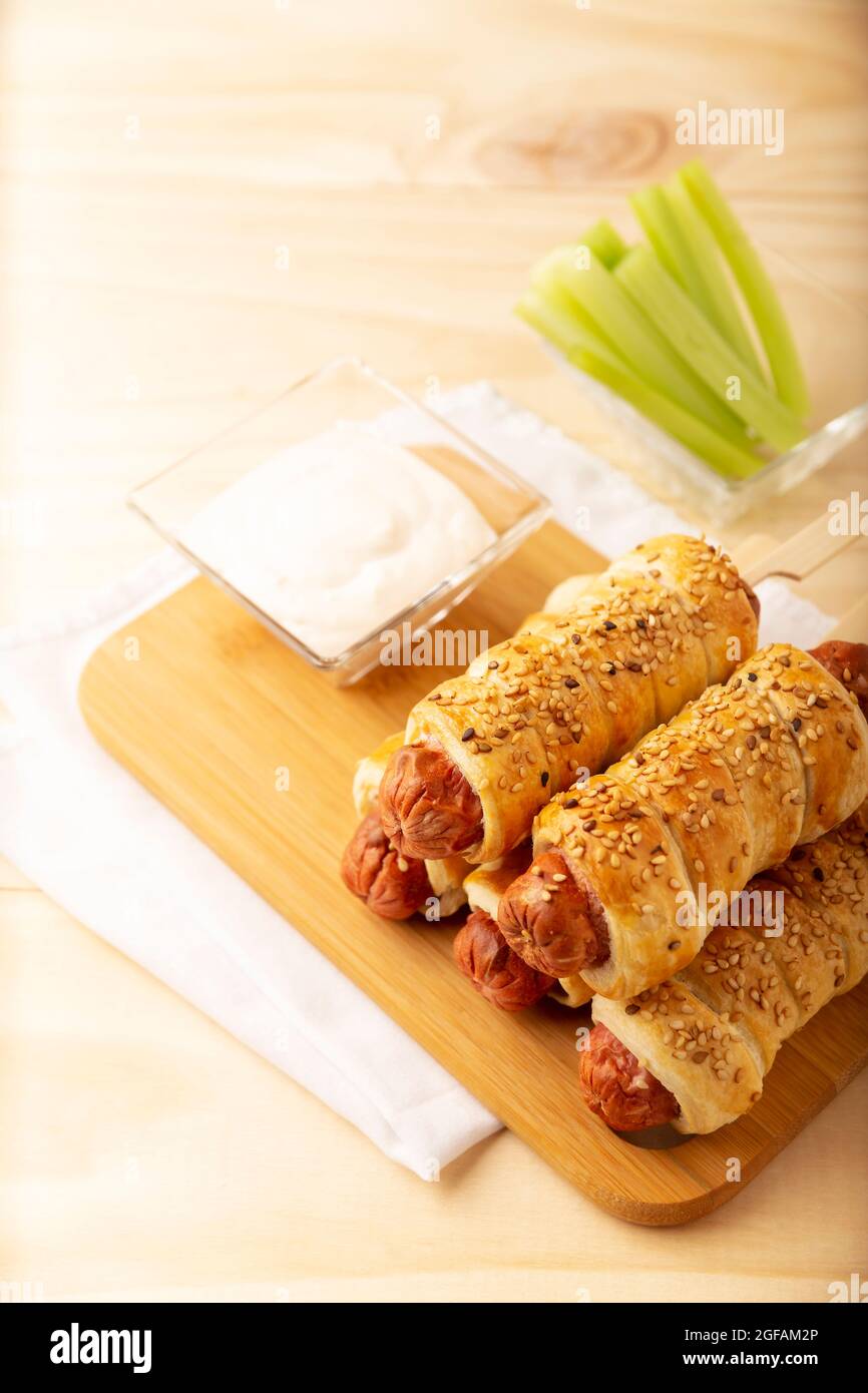 Köstlicher Snack mit Würstchen, eingewickelt in Blätterteig mit Sesam, begleitet von Kirschsellerie-Sticks und Sauce. Serviert auf Holzbrett Stockfoto