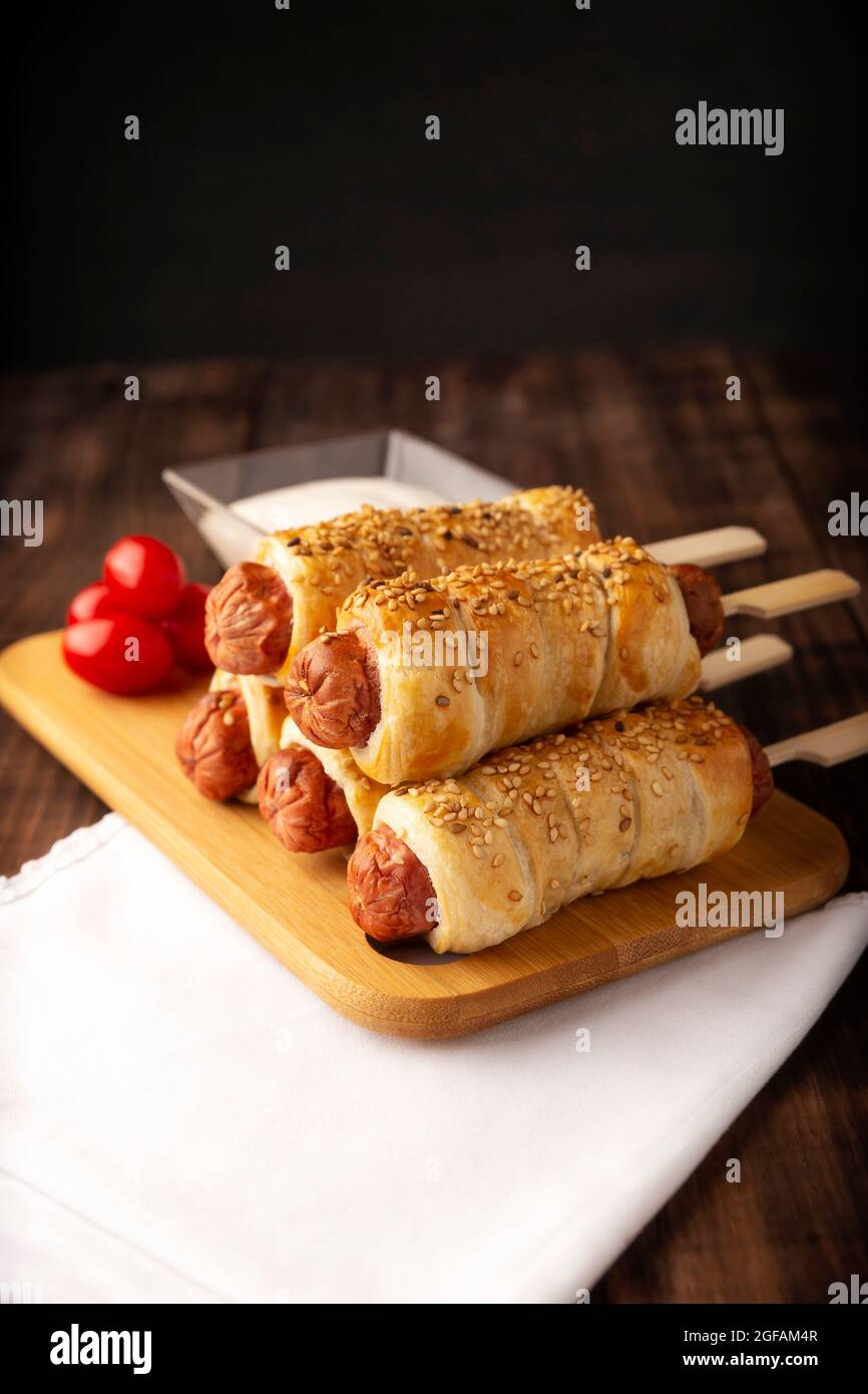 Köstlicher Snack mit Würstchen, eingewickelt in Blätterteig mit Sesam, begleitet von Kirschtomaten und Sauce. Serviert auf Holzbrett Stockfoto