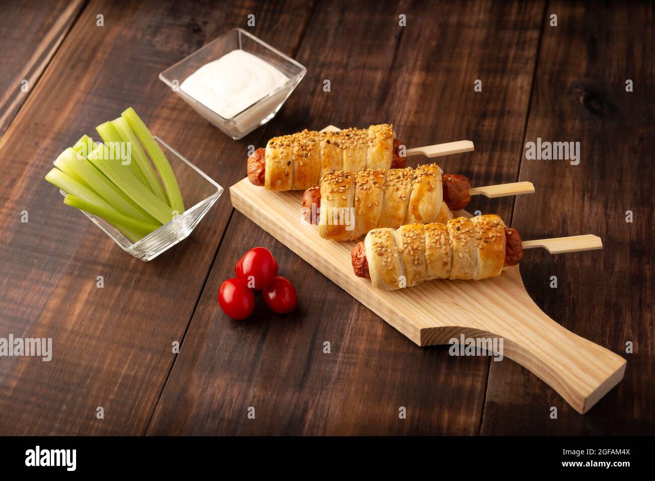 Köstlicher Snack mit Würstchen, eingewickelt in Blätterteig mit Sesam, begleitet von Kirschtomaten, Sellerie-Sticks und Sauce. Serviert auf Holzbrett Stockfoto