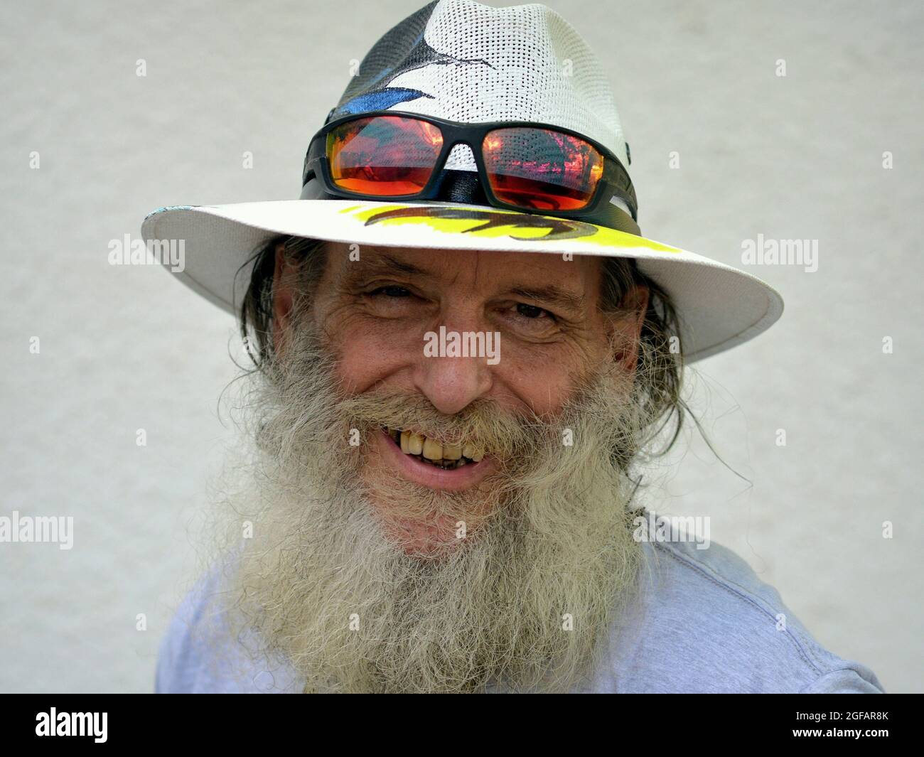 Bärtig optimistischer älterer kaukasischer Mann lächelt und trägt einen bemalten Panama-Hut mit farbenfroher Sonnenbrille am Rand seines Hutes, weißer Hintergrund. Stockfoto