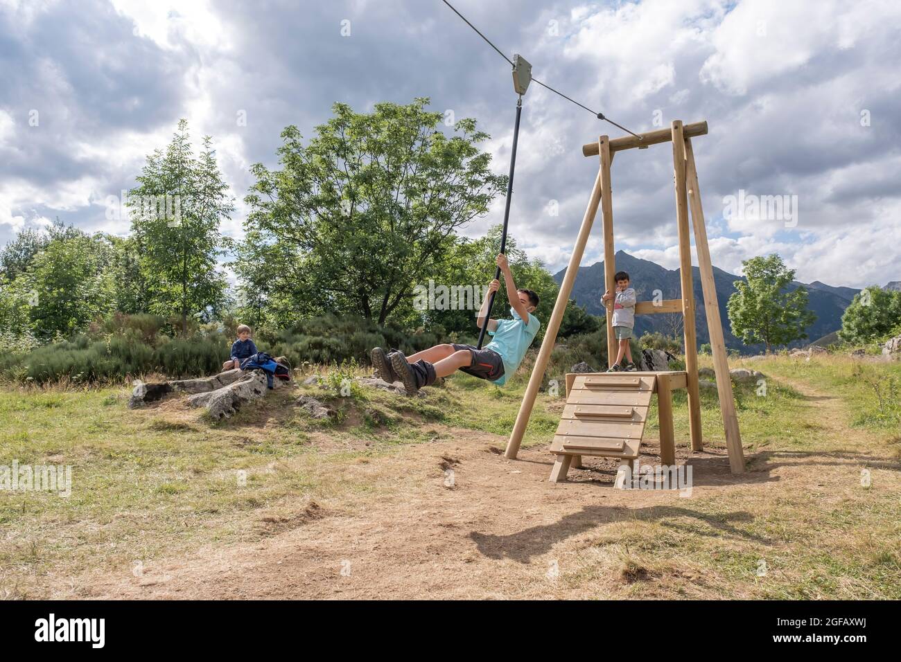 Der Junge mit einer Gesichtsmaske springt im Sommerlager von einer Reißleine auf dem Spielplatz, während andere ihn horizontal angucken Stockfoto