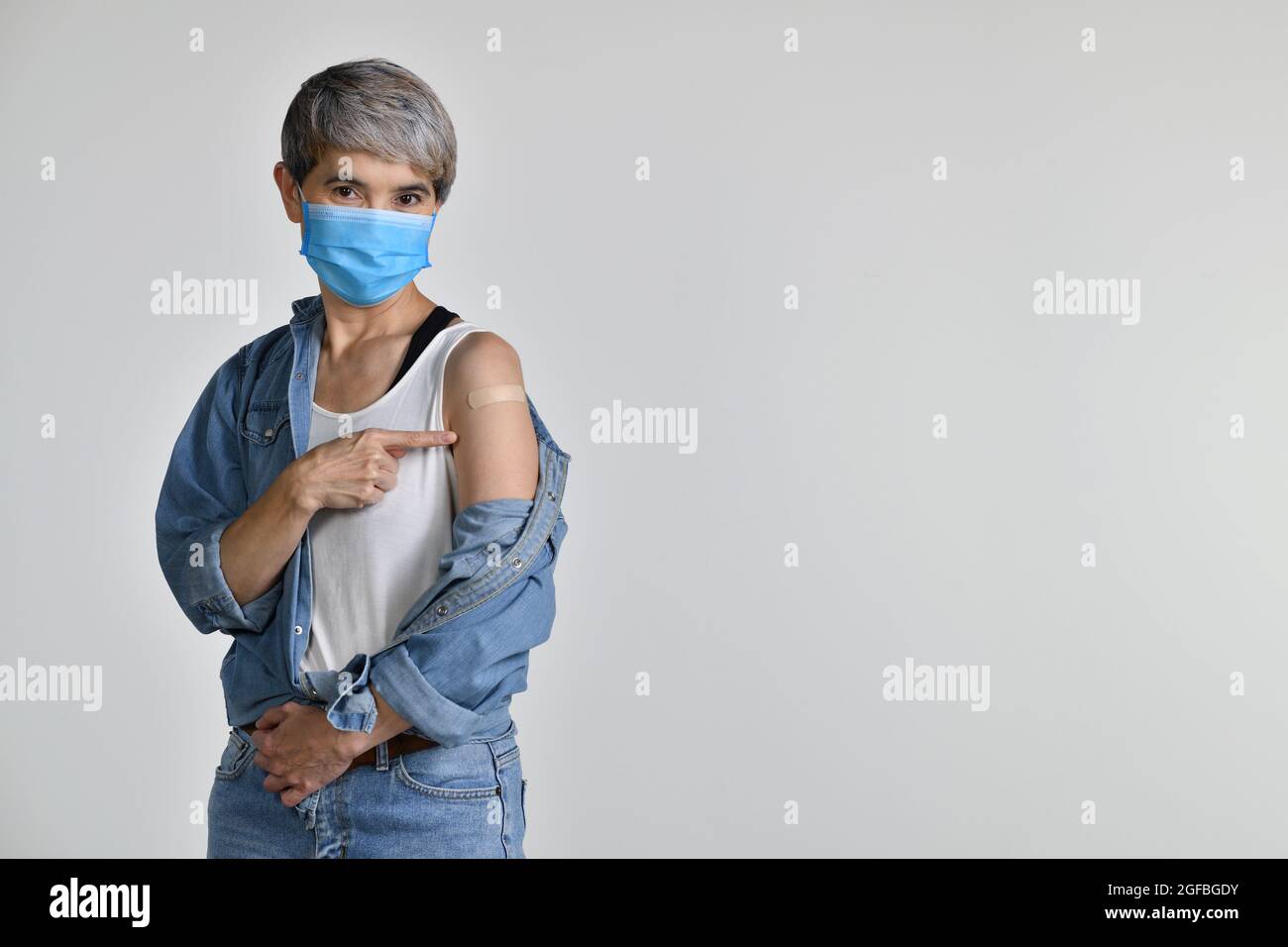 Glückliche Asiatin mittleren Alters geimpfte den Zeigefinger auf die klebende Gipsbandage auf ihrem Arm nach einer antiviralen Injektion des Coronavirus, die auf dem Whit isoliert wurde Stockfoto