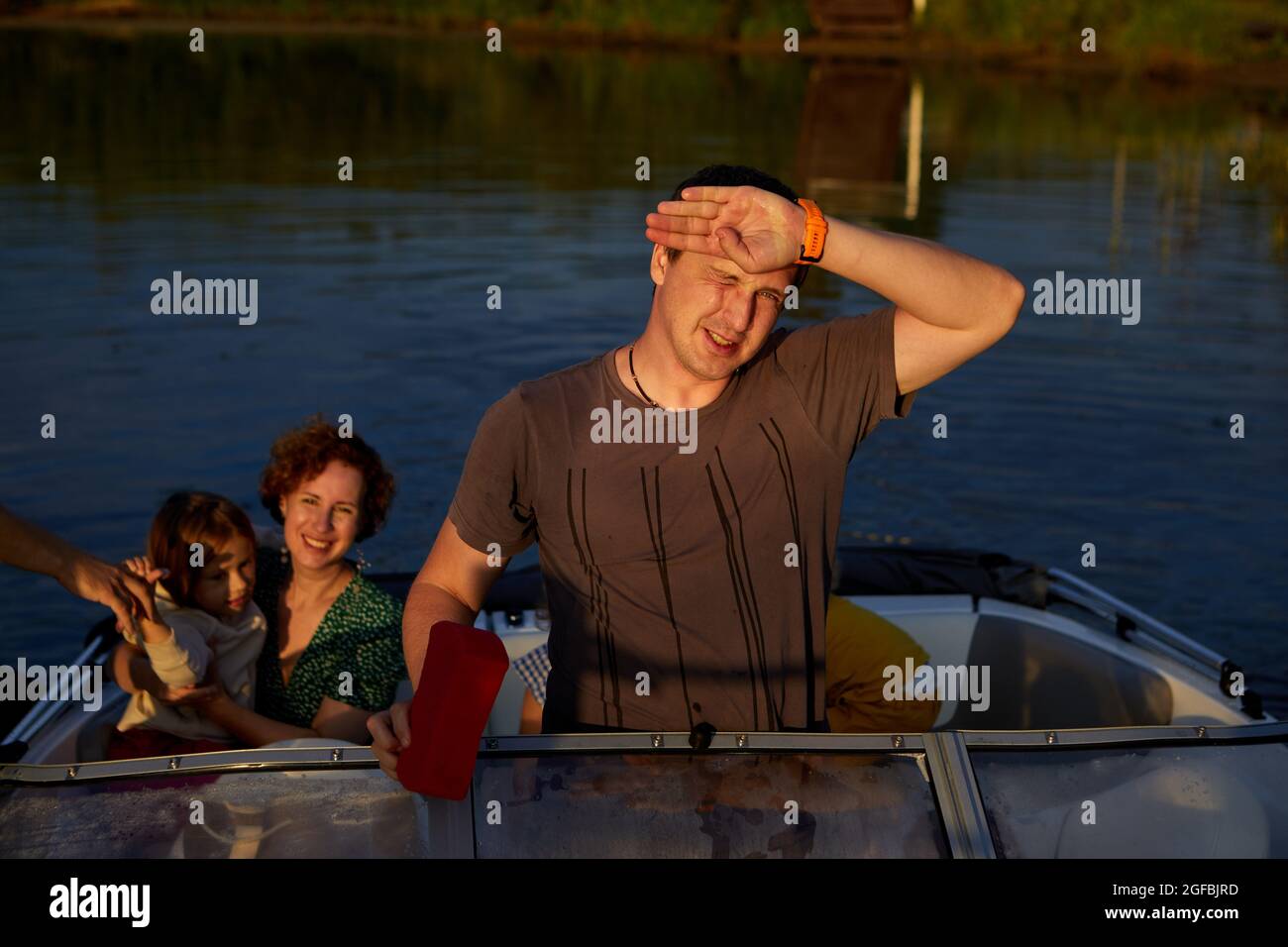 Erwachsener Mann schützt das Gesicht vor Sonnenlicht, während er am Sommertag mit der Familie auf dem See mit dem Motorboot fährt Stockfoto