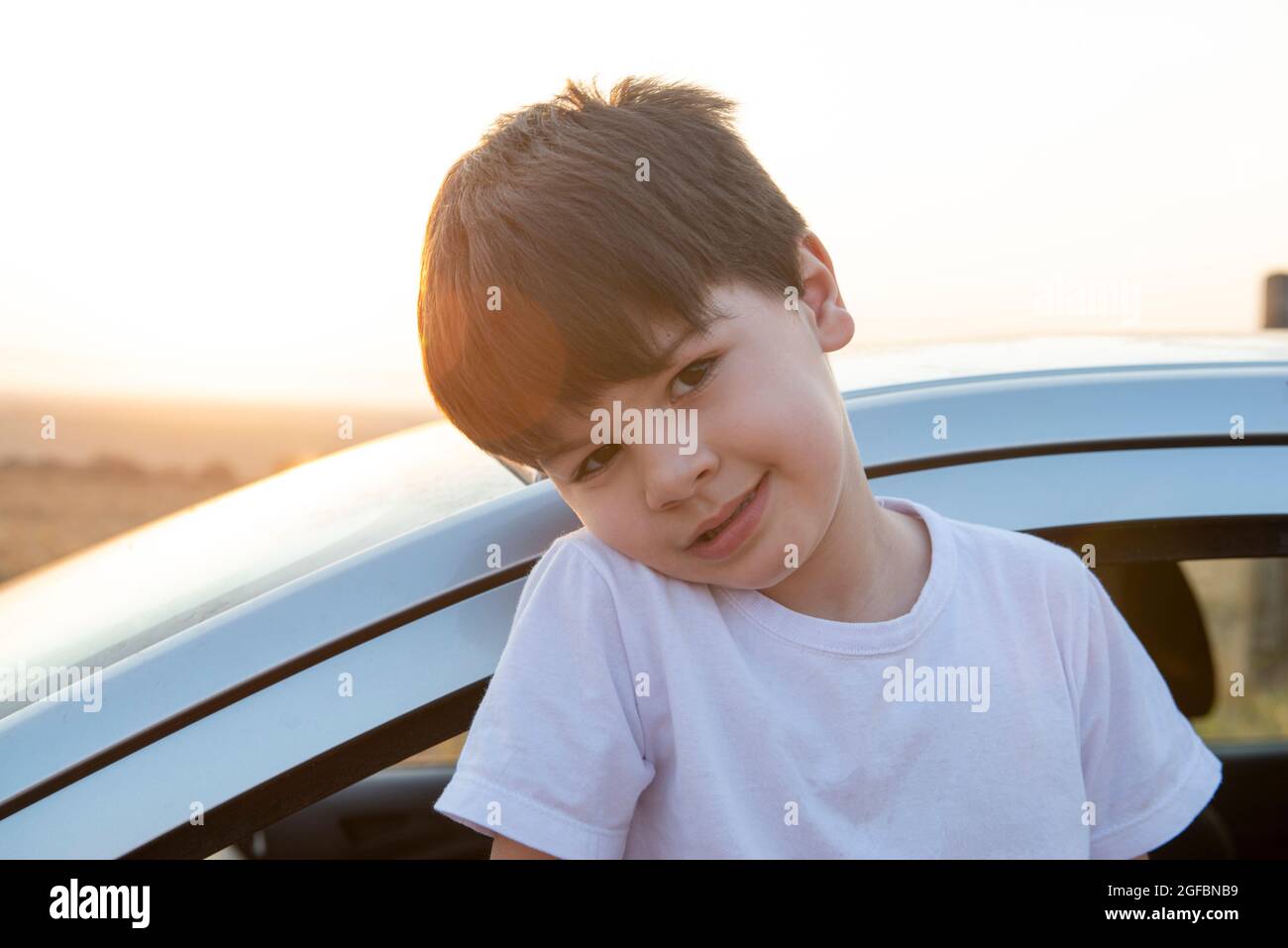 Nahaufnahme eines charmanten kaukasischen Jungen mit schüchternem Lächeln, dessen Körper bei Sonnenuntergang aus dem Auto mit der Sonne im Hintergrund kommt. Stockfoto