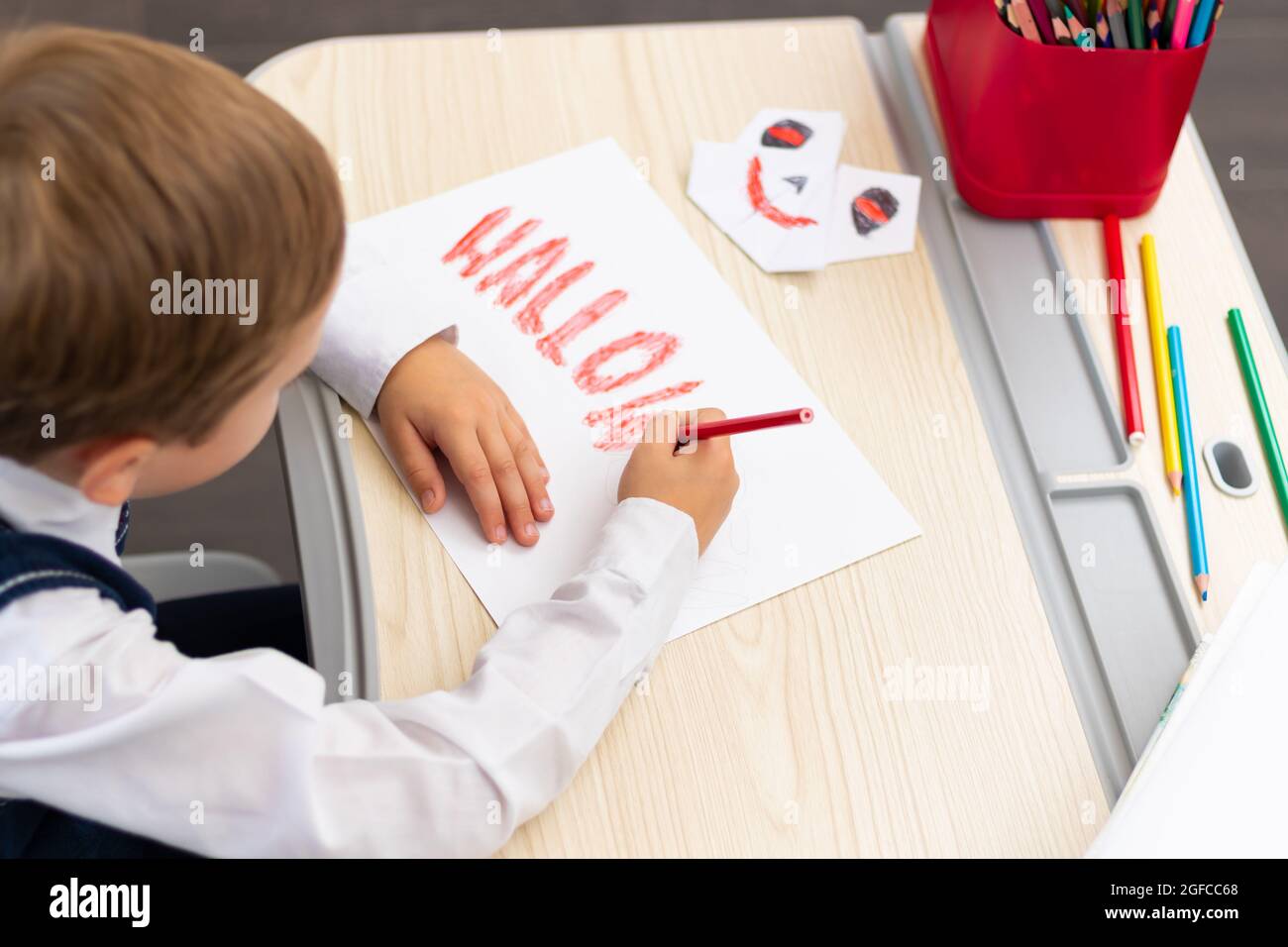 Ein Erstklässler zeichnet mit einem roten, hellen Bleistift auf ein weißes Blatt Papier eine Postkarte für den Feiertag von halloween, während er an einem weißen Tisch sitzt. Se Stockfoto