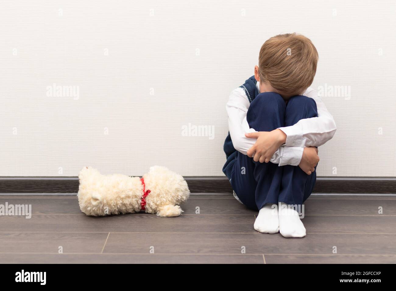 Ein kleiner Junge von sechs ist beleidigt und sitzt auf dem Boden gegen eine weiße Wand neben einem weichen Spielzeug mit geschlossenen Händen. Selektiver Fokus. Nahaufnahme Stockfoto