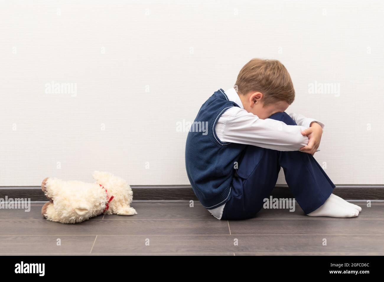 Ein kleiner Junge von sechs ist beleidigt und sitzt auf dem Boden gegen eine weiße Wand neben einem weichen Spielzeug mit geschlossenen Händen. Selektiver Fokus. Nahaufnahme Stockfoto