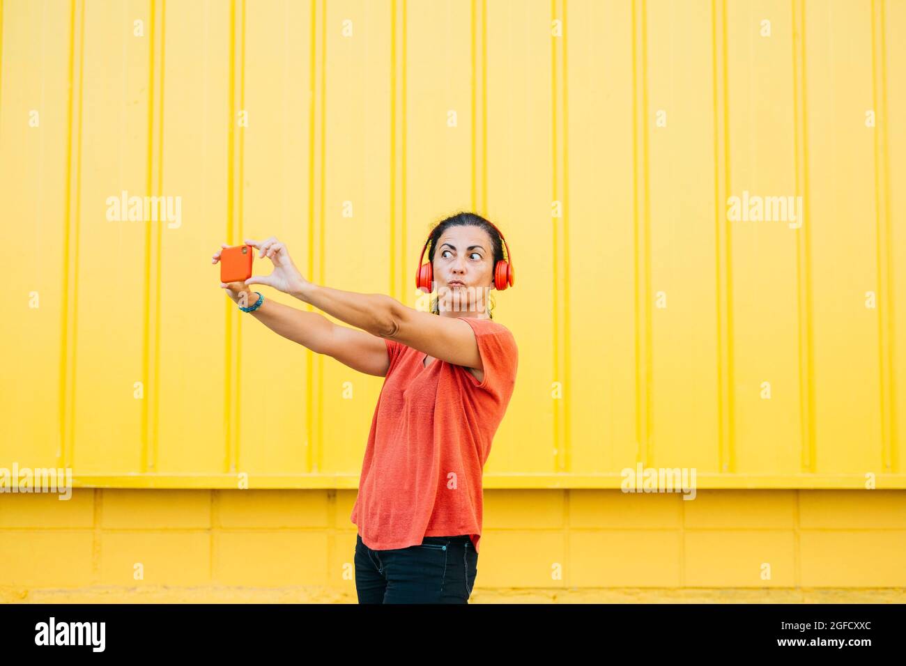 Frau mittleren Alters mit roten Kopfhörern und roten Kleidern, die mit einem roten Smartphone vor gelbem Hintergrund ein Selfie machen Stockfoto