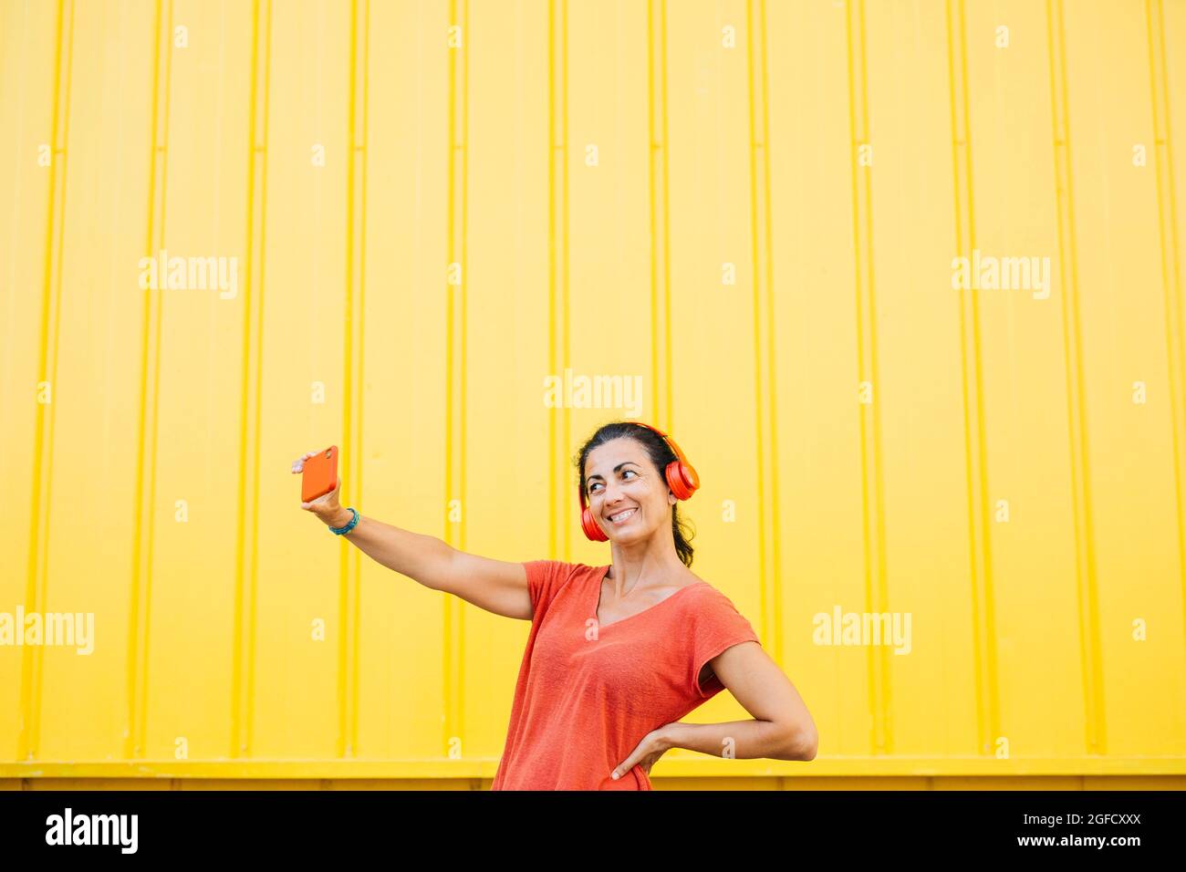 Frau mittleren Alters mit roten Kopfhörern und roten Kleidern, die mit einem roten Smartphone vor gelbem Hintergrund ein Selfie machen Stockfoto