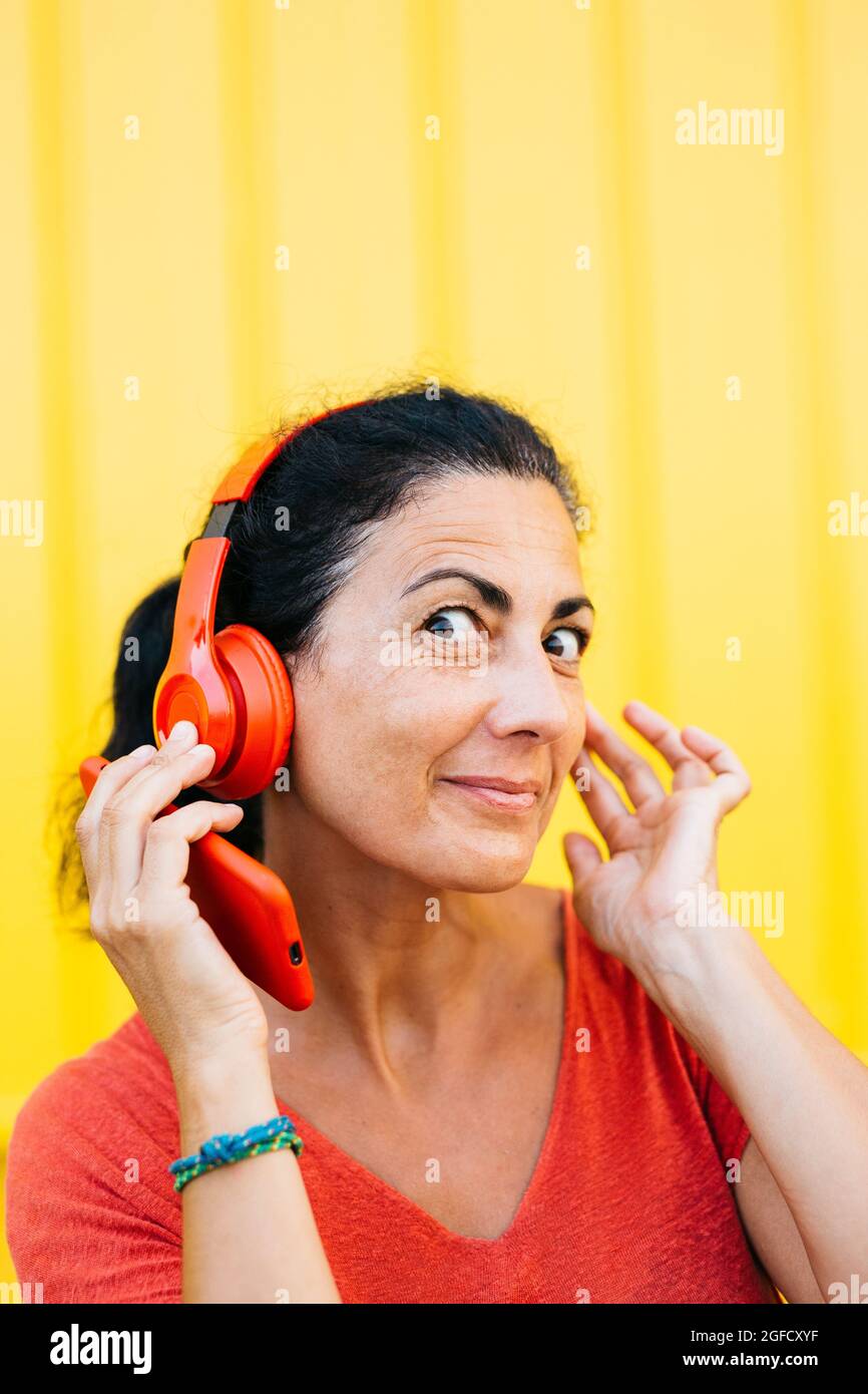 Frau mittleren Alters, die rote Kleidung trägt, Musik mit einem roten Smartphone und einem kabellosen roten Kopfhörer vor einem gelben Hintergrund hört Stockfoto
