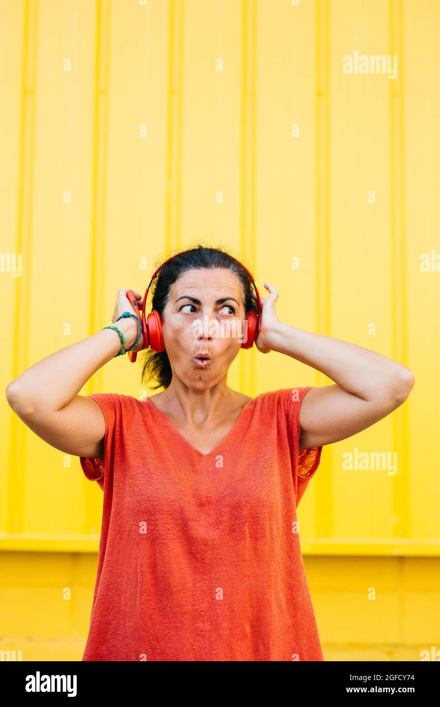 Frau mittleren Alters, die rote Kleidung trägt, Musik mit einem roten Smartphone und einem kabellosen roten Kopfhörer vor einem gelben Hintergrund hört Stockfoto