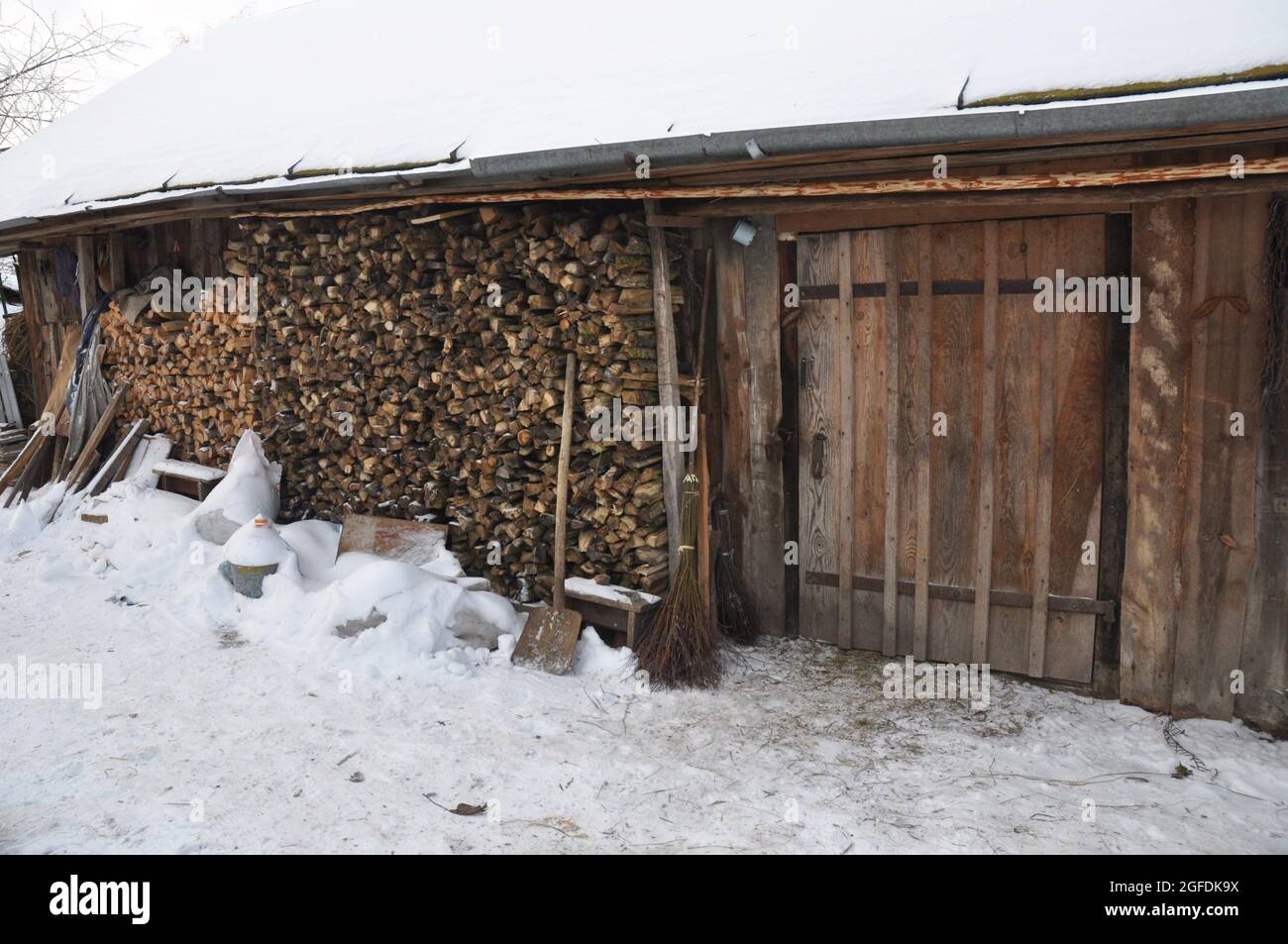 Eine alte rustikale ländliche Holzscheune mit einem Dach bedeckt mit Schnee und Feuerholz entlang der Scheunenwand im Winter gestapelt. Gepflegter Holzstapel an der Scheune. Stockfoto