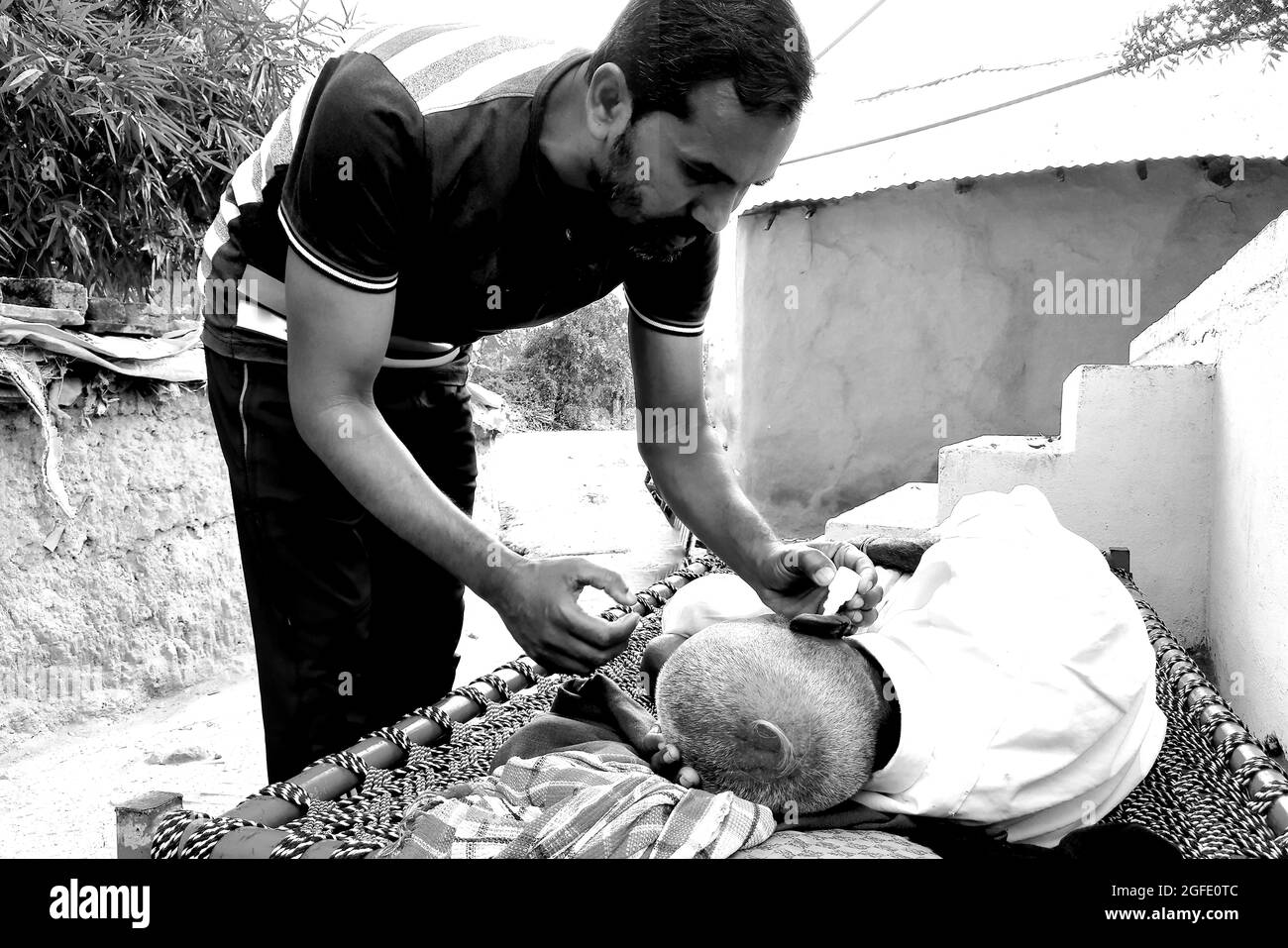 Ein ländlicher Jugendlicher, der seinem kränkelnden Großvater Medizin ins Ohr gießt, ländliche Umgebung. Schwarzweiß-Bild. Stockfoto