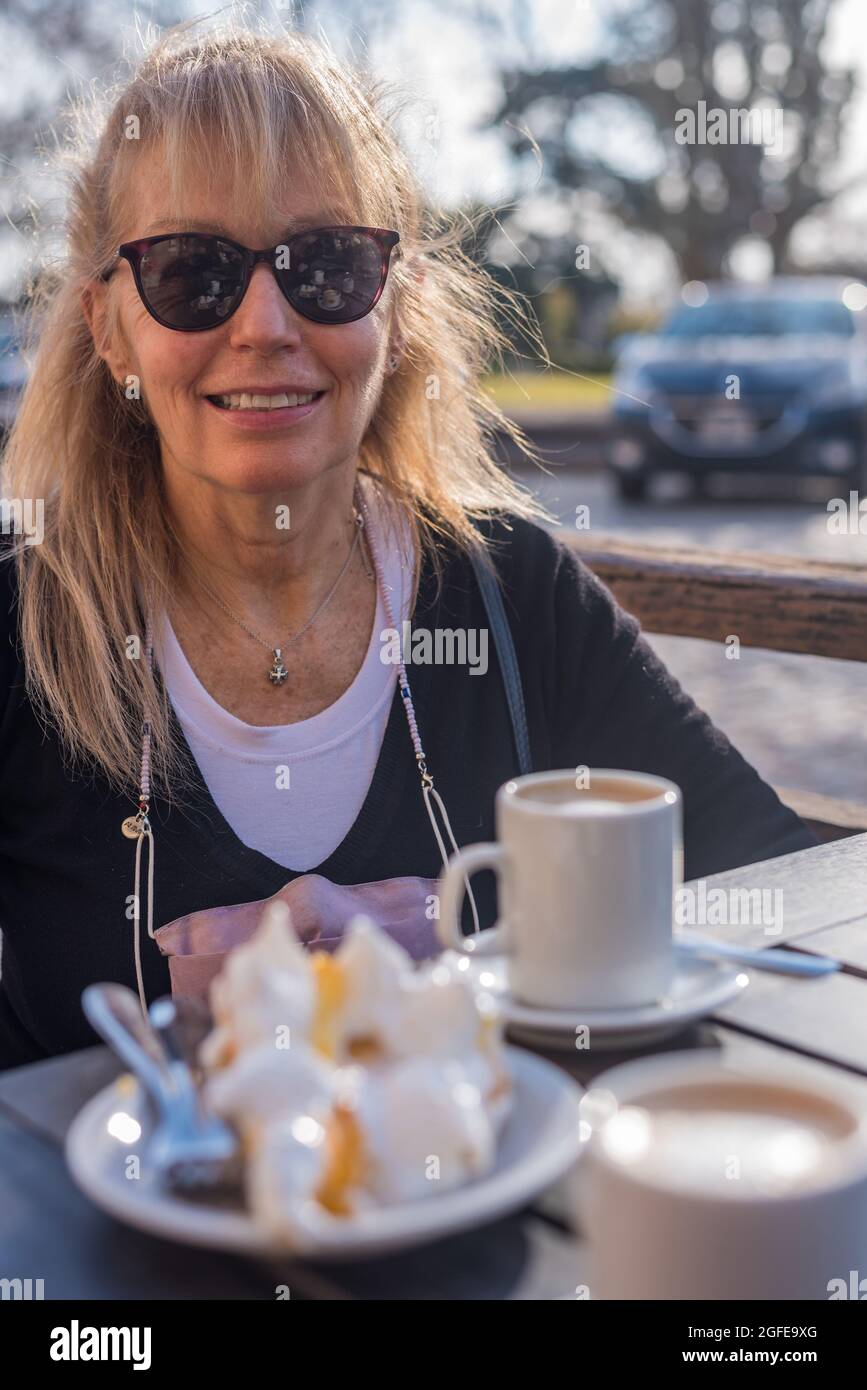 Ältere Erwachsene Frau, die lächelt und eine Brille mit Latte und Zitronenkuchen trägt. Blick auf die Kamera Stockfoto