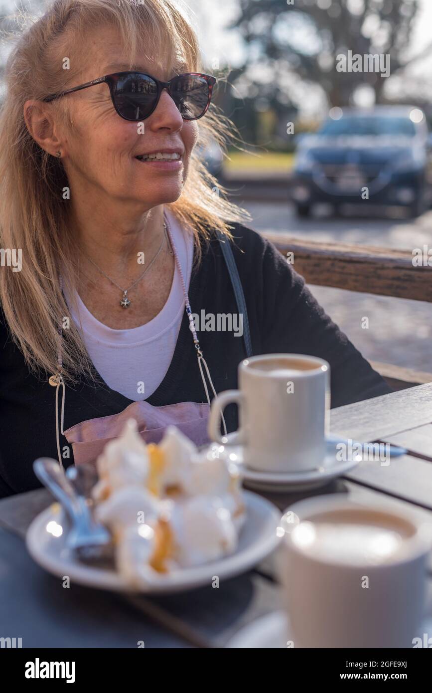 Ältere Erwachsene Frau, die lächelt und eine Brille mit Latte und Zitronenkuchen trägt. Wegschauen Stockfoto
