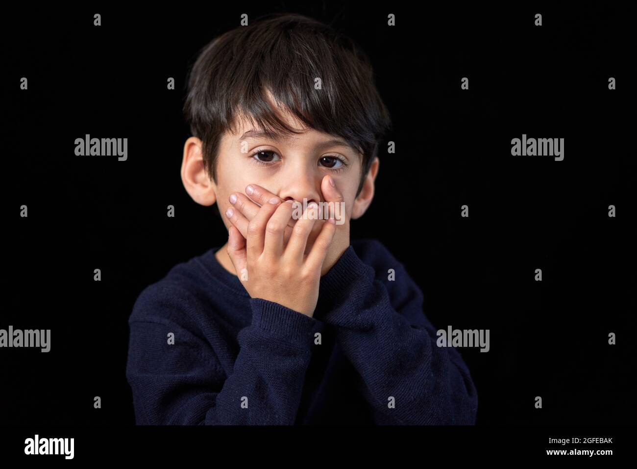 studio-Porträt eines ernsthaften lateinischen Jungen, der die Kamera betrachtet, die seinen Mund mit beiden Händen bedeckt. Schwarzer Hintergrund und Kopierraum. Konzept der Stille oder Stockfoto