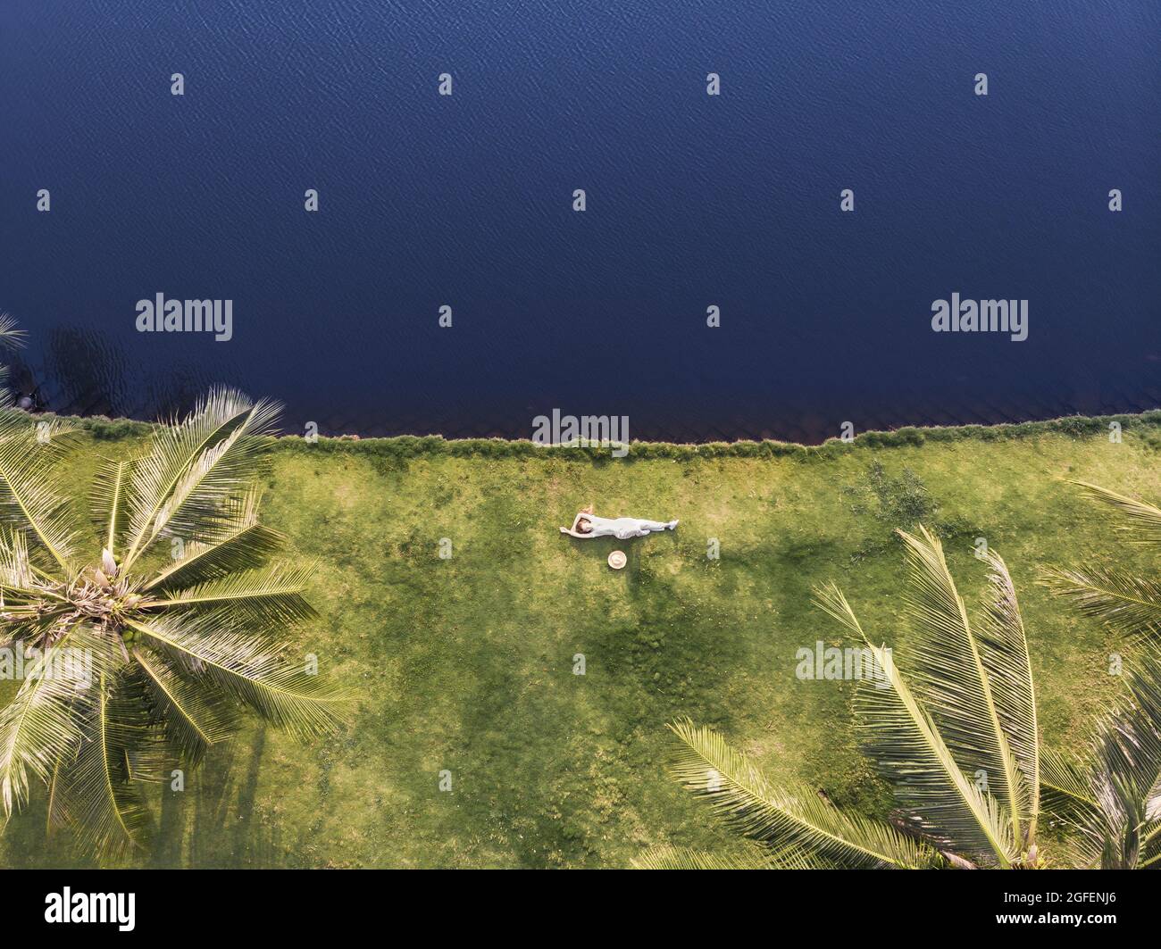 Kaukasische junge Frau in weißem Kleid, die auf dem Gras am Ufer zwischen zwei Palmen liegt. Foto auf der Seite oben. Bild von der Drohne Stockfoto