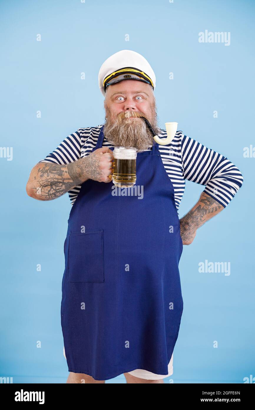 Positiv fetter Mann im Matrosenanzug mit Schürze hält Craft-Bier auf hellblauem Hintergrund Stockfoto