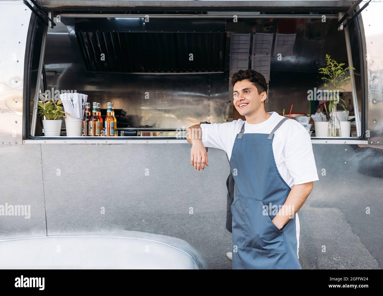 Junger Geschäftsinhaber in einer Schürze. Verkäufer, der sich auf einen Food Truck stützt. Stockfoto