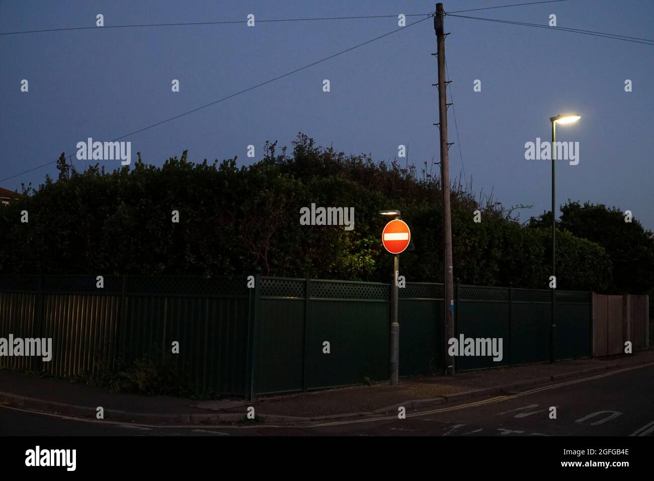 East Wittering, Großbritannien, 25. August 2021: In der Abenddämmerung ragt ein beleuchtetes No Entry-Schild scharf gegen eine hohe Hecke an einer Vorstadtstraße heraus. Stockfoto