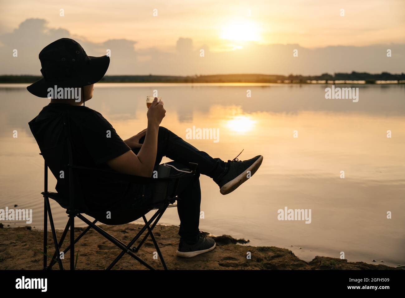 Asiatischer junger Mann, der in einem Touristenstuhl sitzt und eine Tasse mit Tee oder Kaffee hält, den Blick auf die Landschaft des Flusses bei Sonnenuntergang genießt. Tra Stockfoto