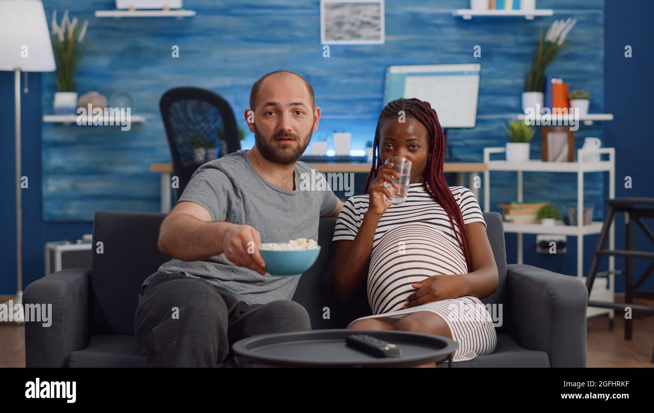 POV der interracial Familie Blick auf Kamera auf der Couch sitzen. Gemischtes Rennpaar erwartet Kind beim Fernsehen. Kaukasischer Mann bringt Glas Wasser für schwarze Schwangere Stockfoto