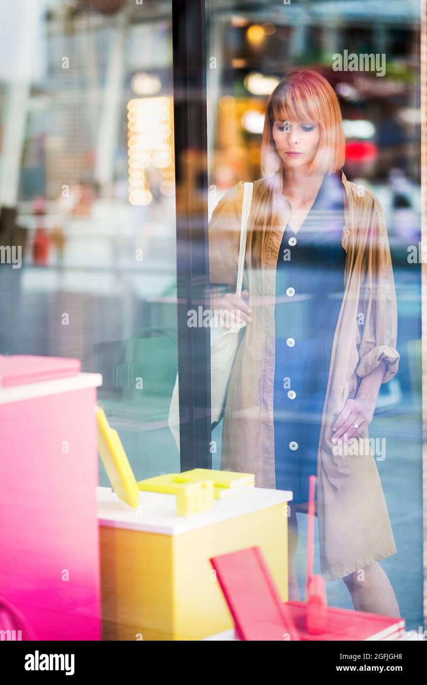 Elegante Frau beim Schaufensterbummel in einer Stadtstraße, die vom Inneren des Geschäfts aus durch das Glas betrachtet wird, während sie die Waren betrachtet, die ausgestellt werden Stockfoto