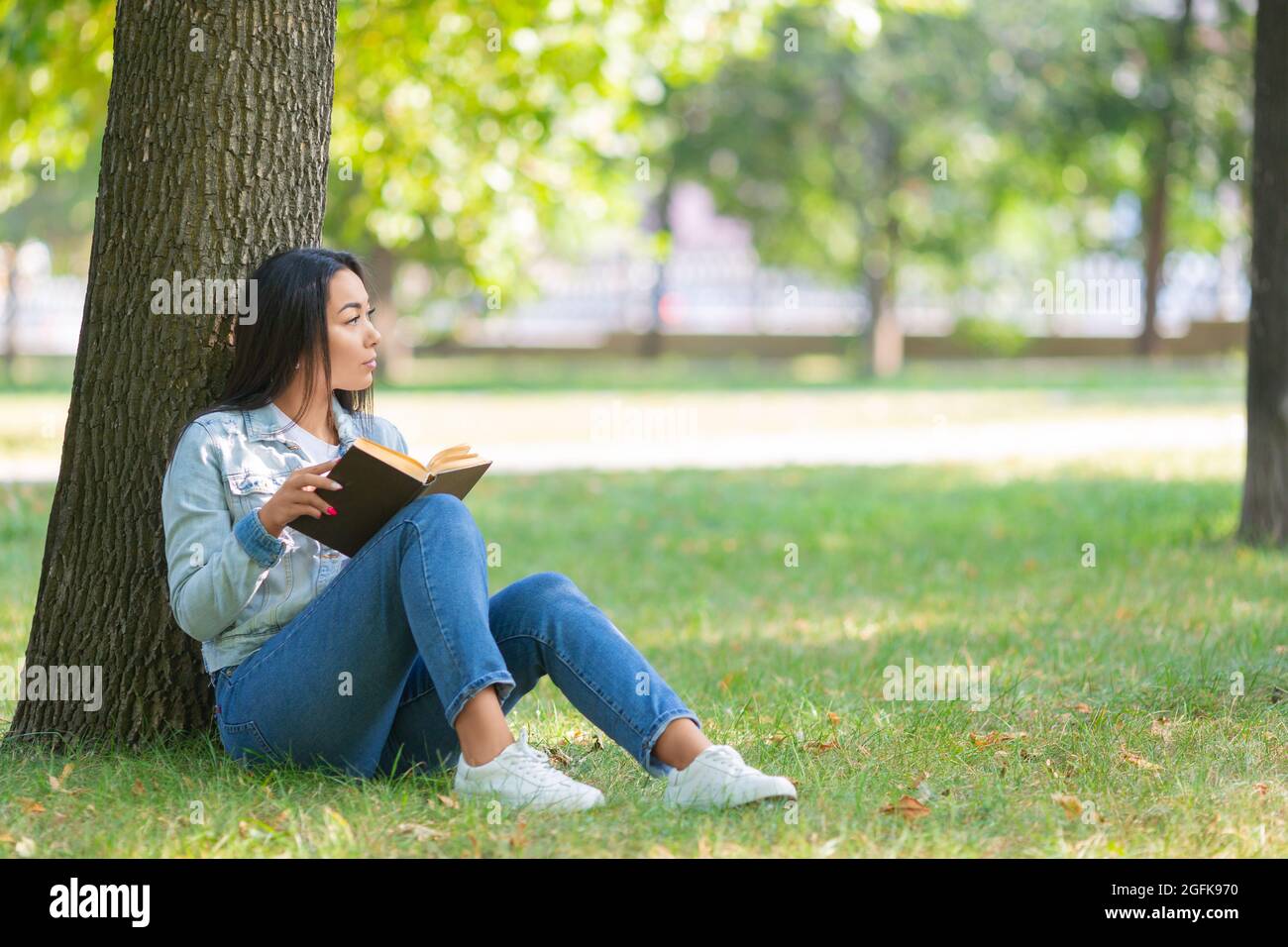 Asiatische Frau mit einem Buch im Park. Sie sitzt auf dem Gras im Park und schaut nebeneinander. Das Konzept der Ruhe von Gadgets, Smartphones und der Inte Stockfoto