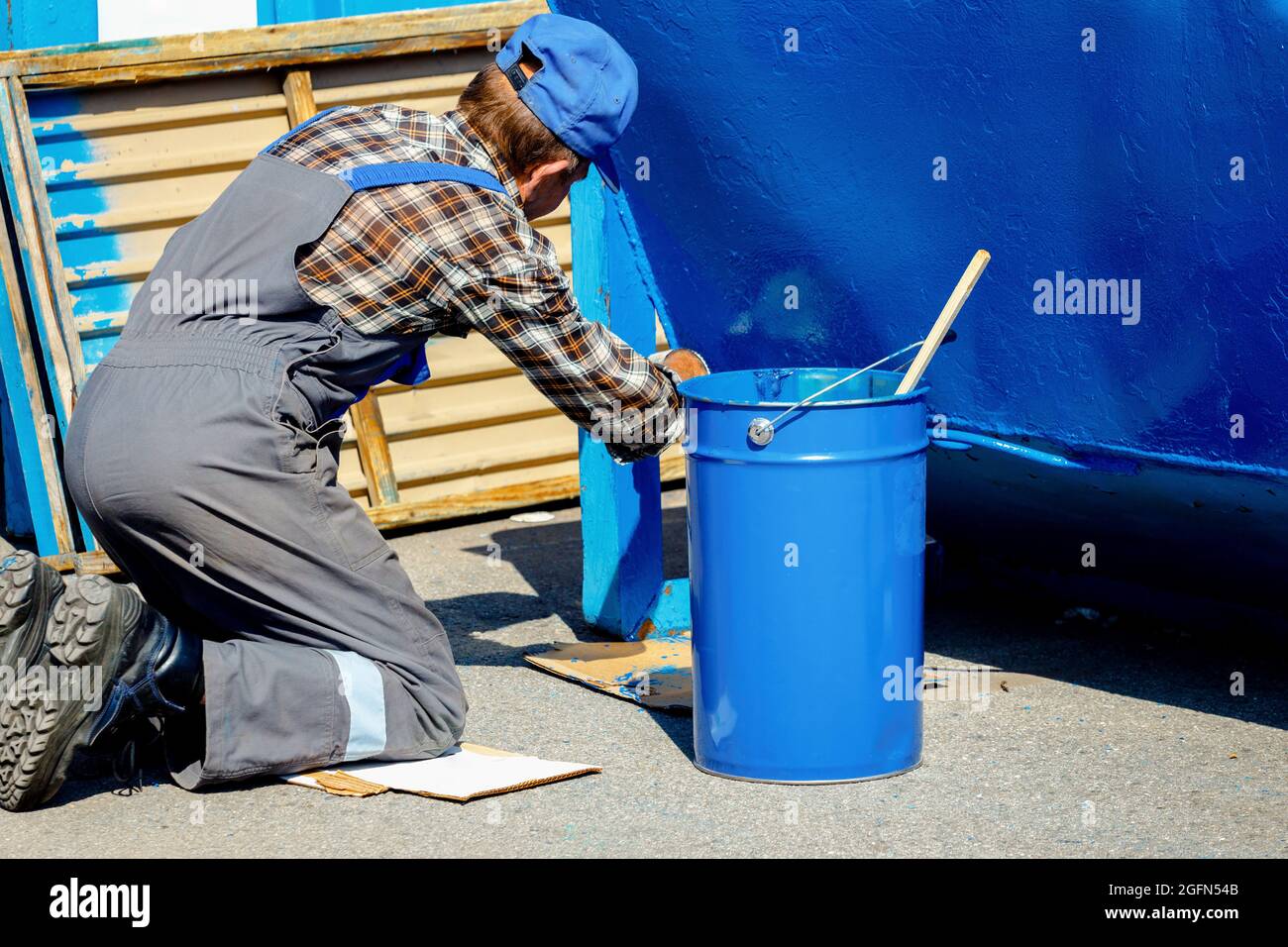 Ein alter Mann in einem Arbeitsanzug malt mit einem Pinsel auf der Straße eine Mülltonne oder einen Behälter. Teilzeitarbeit für einen Rentner. Mühsal. Stockfoto