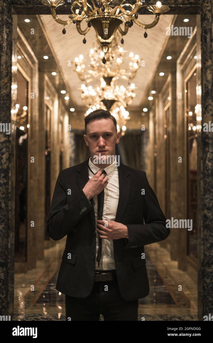 Porträt eines jungen, gutaussehenden Geschäftsmannes in elegantem Geschäftsanzug, der mit schönen teuren Kronleuchtern auf dem Flur herumläuft Stockfoto