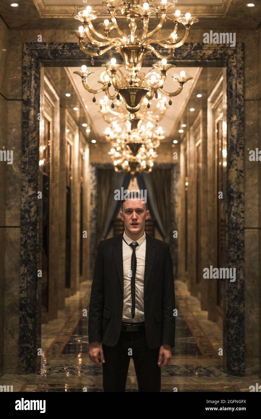 Porträt eines jungen, gutaussehenden Geschäftsmanns in elegantem Geschäftsanzug, der mit schönen teuren Kronleuchtern auf dem Flur herumläuft. Ein reiches Gebäude. Stockfoto