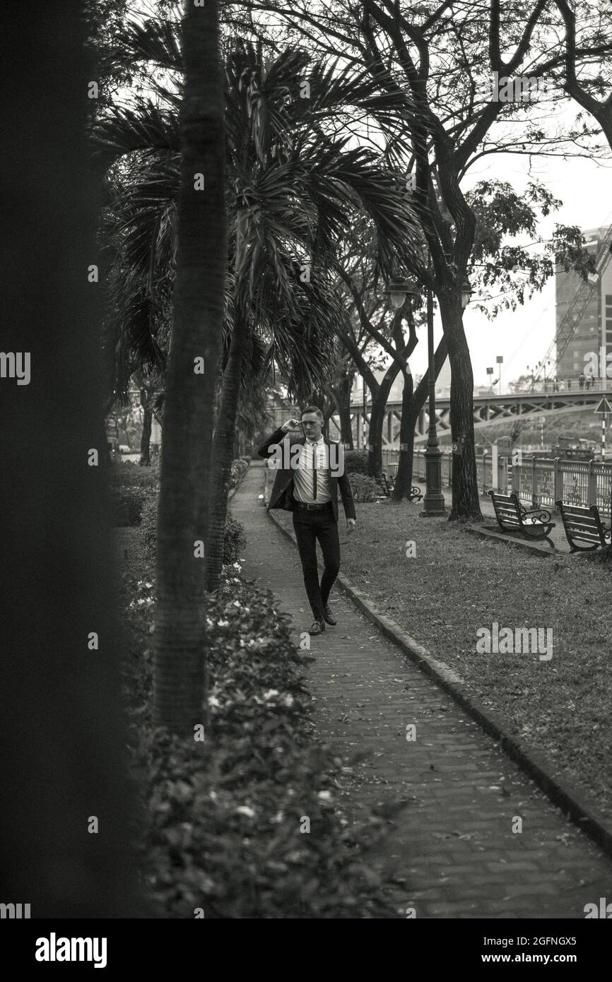 Hübscher junger Kaukasier in schwarzem Anzug und Sonnenbrille, der im Park spazieren geht. Park mit tropischen Pflanzen. Schwarzweiß-Foto Stockfoto