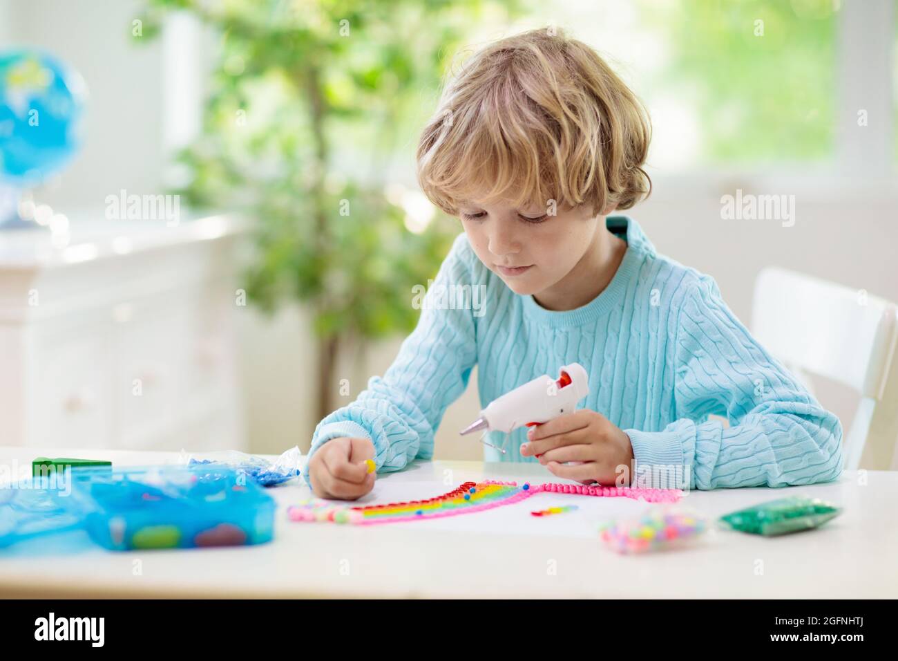 Basteln für Kinder. Kind mit Heißklebepistole. Kreativer kleiner Junge, der Knöpfe und Perlen im Regenbogen-Design klebt. Kunst und Kreativität Klasse in der Schule Stockfoto