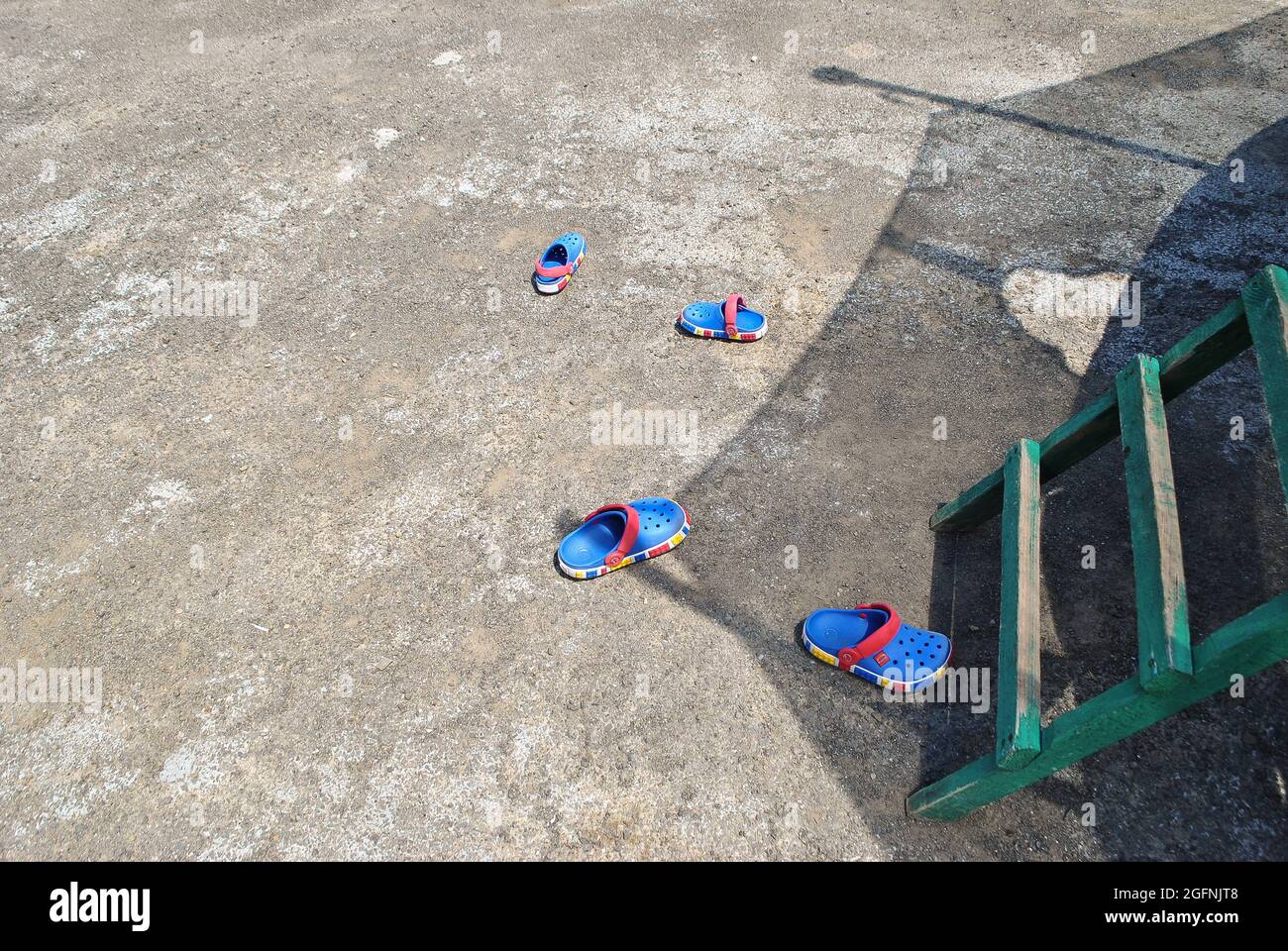 In der Nähe einer Holztreppe auf einem Trampolin auf einem Betonboden sind zwei Paar farbige Kindersandalen verstreut Stockfoto