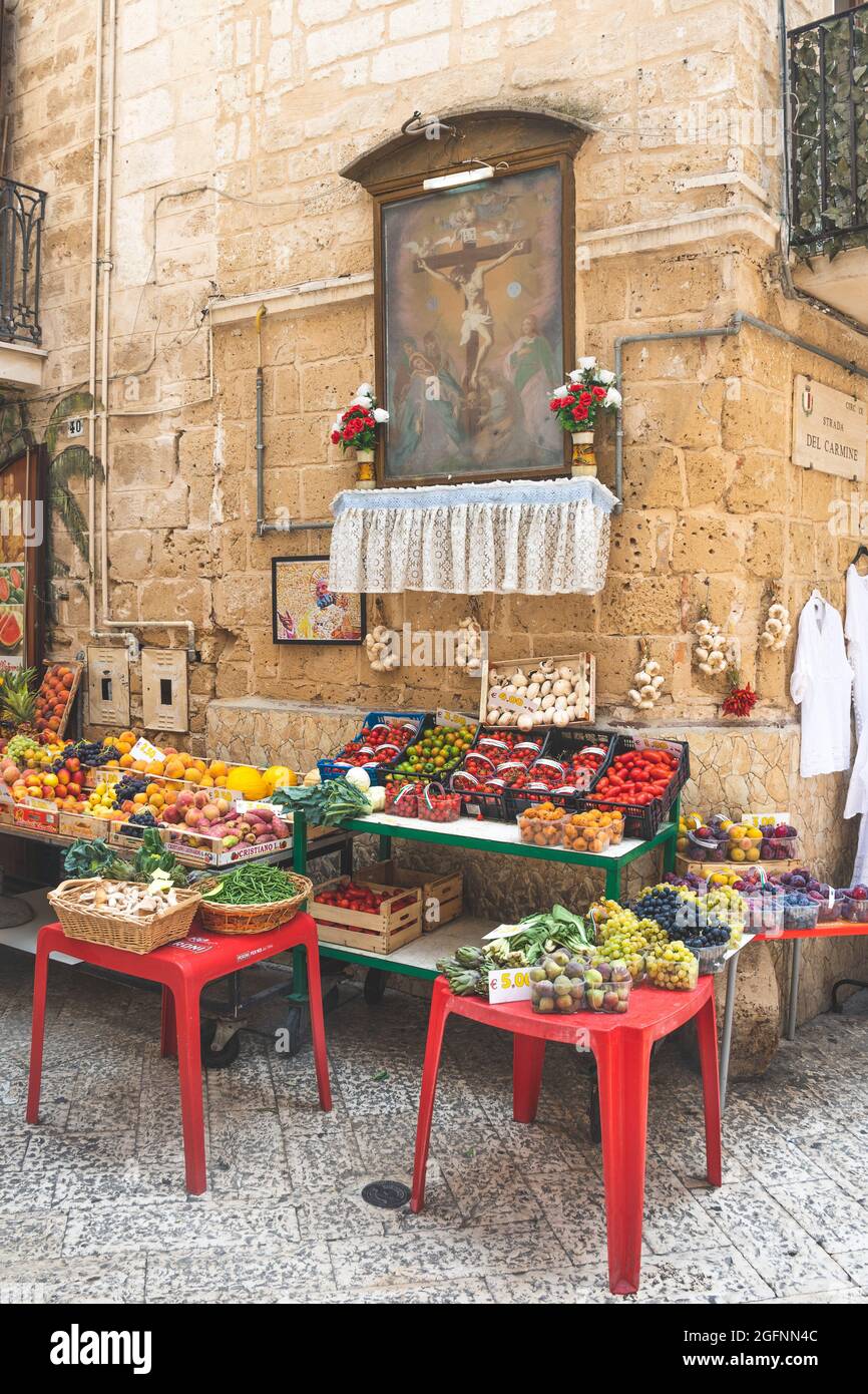 Blick auf eine schmale Straße in der Altstadt von Bari, Apulien, Italien, traditioneller offener Obstmarkt mit religiösen Bildern und verschiedenen lokalen Obst- und Gemüsesorten Stockfoto
