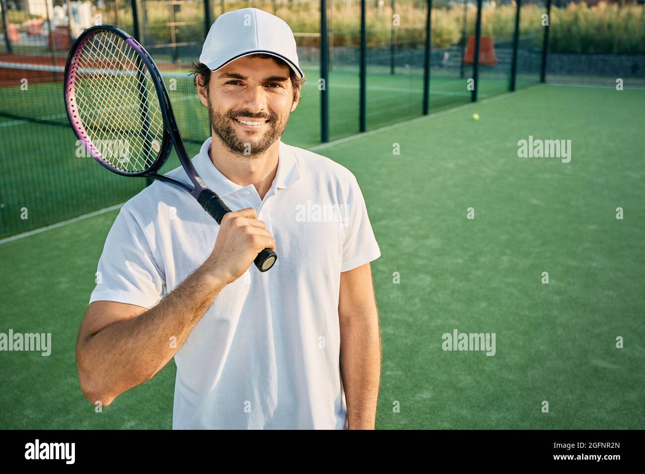 Porträt eines hübschen Tennisspielers mit einem Schläger in der Hand, der in weißer Uniform auf einem Tennisplatz steht Stockfoto