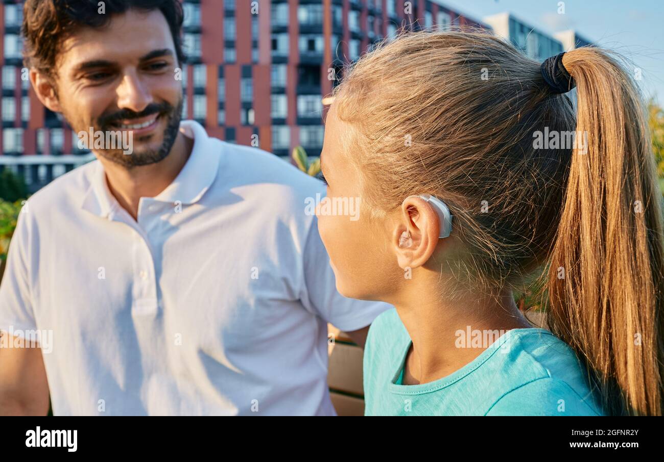 Positives weibliches Kind mit einer Hörbehinderung nutzt ein Hörgerät, um mit ihrem Vater im Freien zu kommunizieren. Hörlösungen Stockfoto